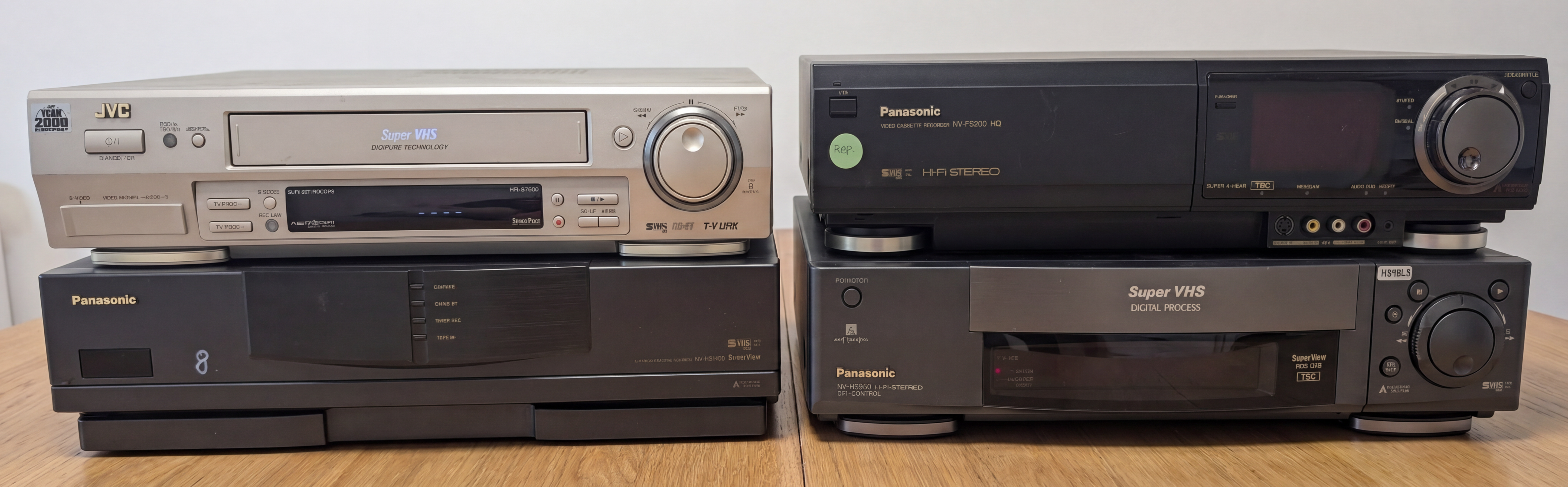Vier Heim-Videorekorder auf einem Holztisch: links ein JVC Super VHS über einem Panasonic, rechts ein Panasonic NV-FS200 HQ über einem Panasonic Super VHS